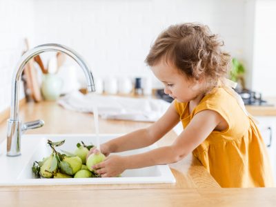 Cute little girl wearing linen mustard dress washing pears and apples in the sink in the white kitchen scandinavian style